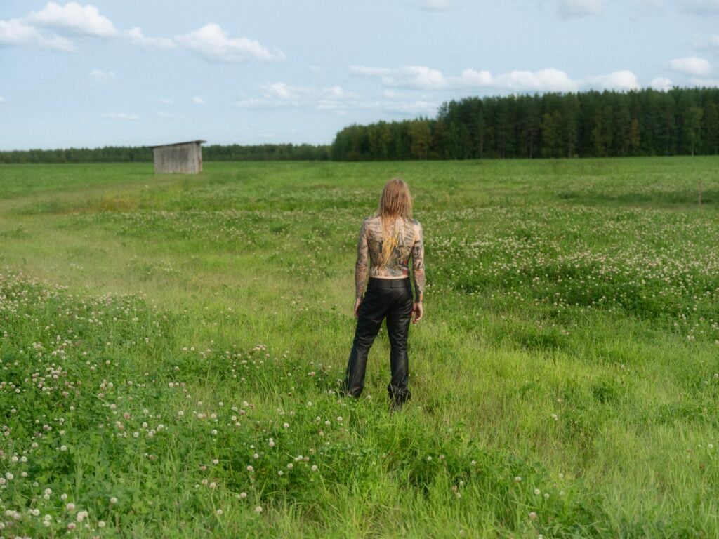 Henkilö seisoo niityllä, taustalla lato ja metsä.